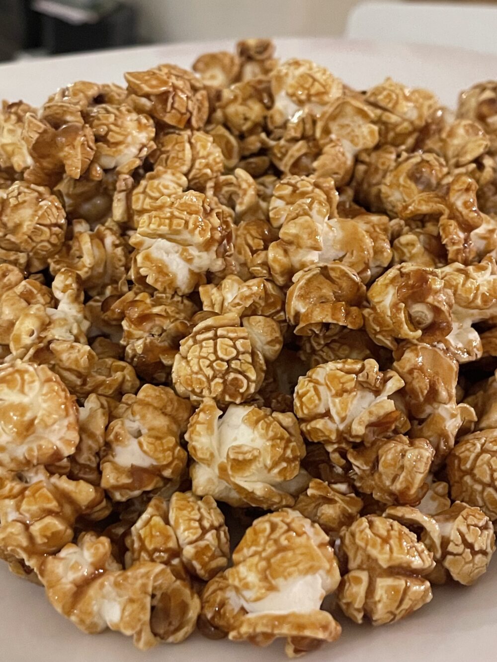 Popcornaa Classic Caramel Popcorn served on a white plate. The golden, caramel-coated kernels glisten under the light, showcasing their crispy texture and rich, caramel coating. The clean, white background highlights the popcorn’s delicious, handcrafted quality."